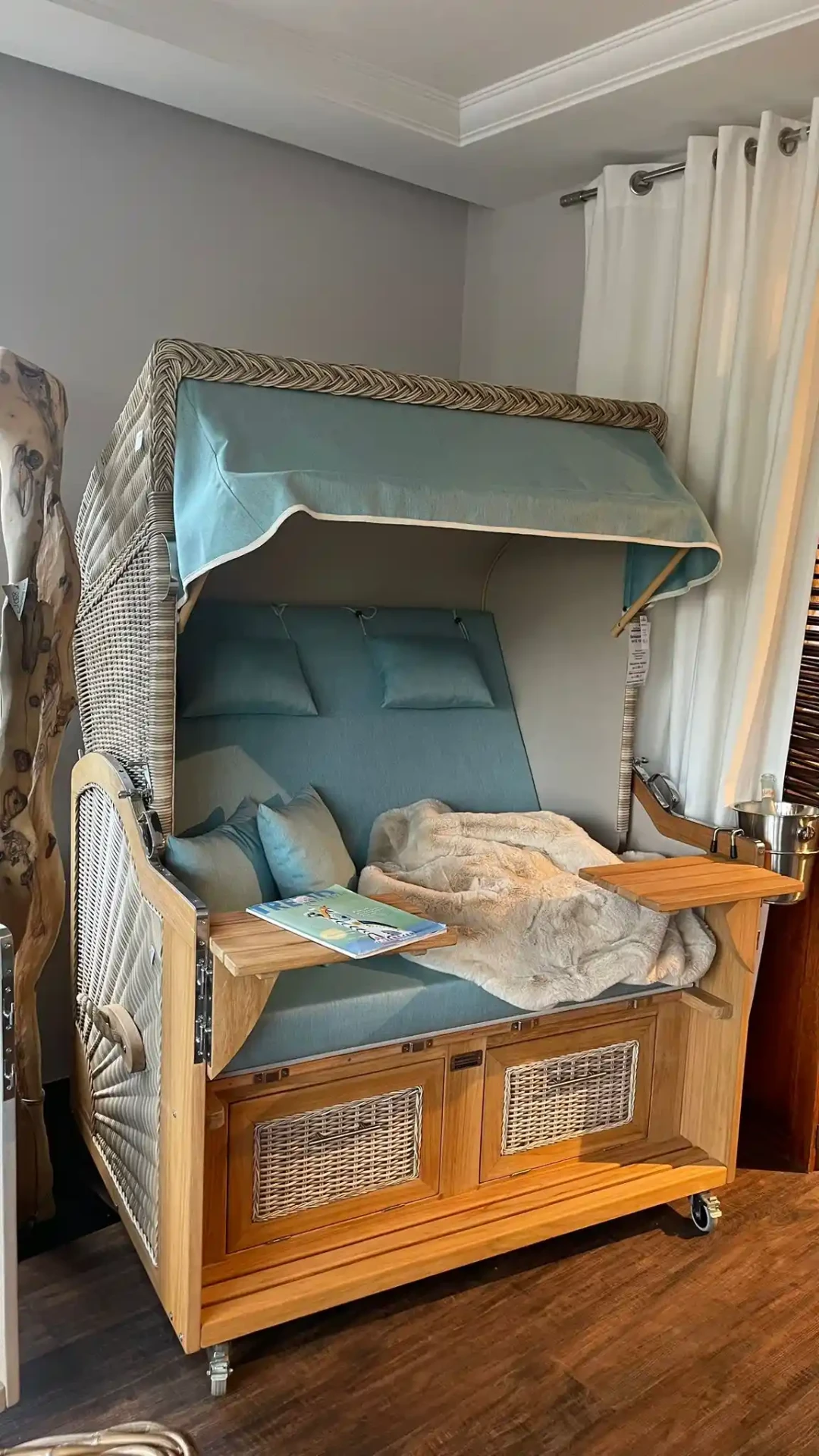 Strandkorb mit türkisfarbenen Polstern, Teakholz-Tischchen und Kuscheldecke im Showroom