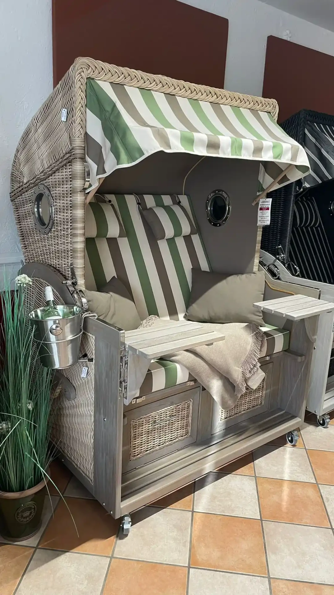Gartenstrandkorb mit grün-beige gestreiften Polstern, Sektkühler und ausklappbaren Tischen